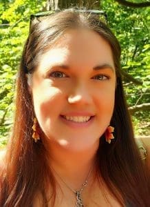 Headshot of Caitlin – UX/UI, Graphic Design, Cultural Design, Writing. A smiling image of a woman in an outdoor setting, with glasses on top of her head, log straight brown hair, and brightly coloured beaded sunflower earrings.